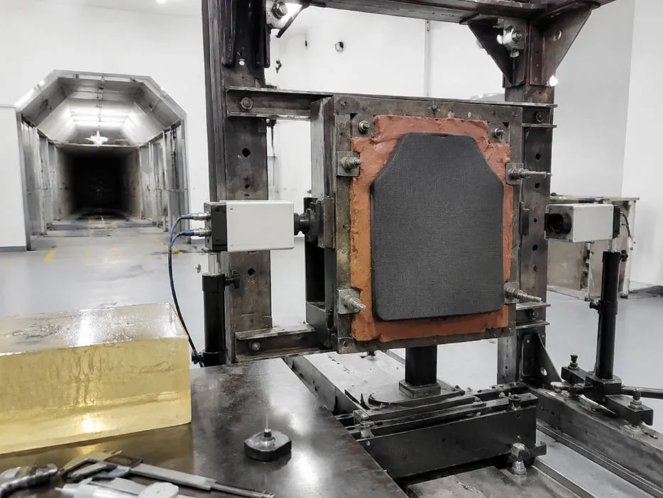 Ballistic testing laboratory with body armor panel mounted on clay backing in automated test rig